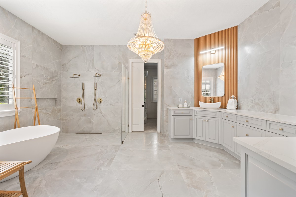 Primary bathroom in large marble tile with a freestanding tub, a crystal chandelier, a double walk-in shower and a wood-slat panel behind the vanity mirror.