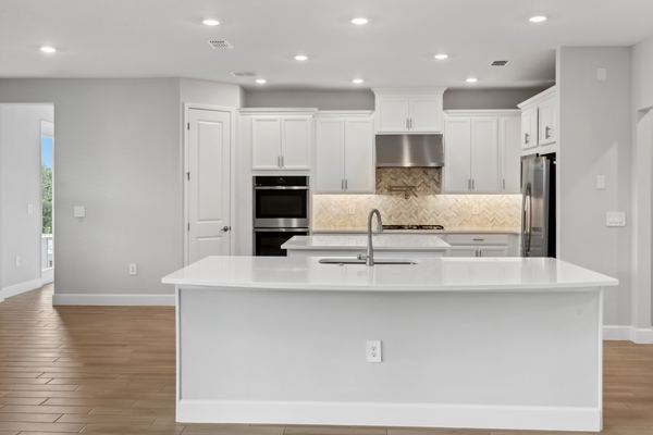 White kitchen with a large quartz island and sink, a herringbone marble backsplash, a stainless chimney hood and double wall ovens.