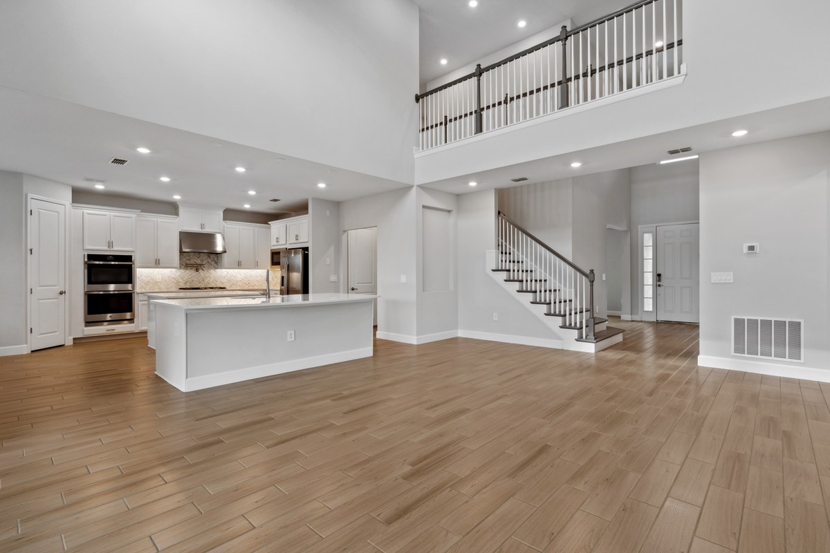 Empty double-height living room in wood-look tile, with an iron-baluster staircase, an upstairs landing and the open kitchen behind.