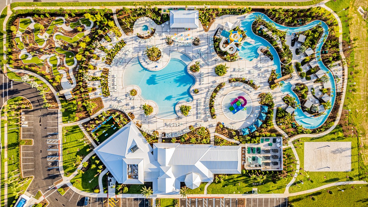 Straight-down aerial of a community water park at golden hour: a free-form pool ringed by loungers and palms, a lazy river curving around it, a splash pad, a mini-golf course and the club house with its parking lot.