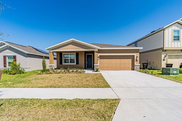 Single-storey home in tan stucco with a gabled entry porch, black shutters, stone corner accents and a two-car garage on a concrete drive.