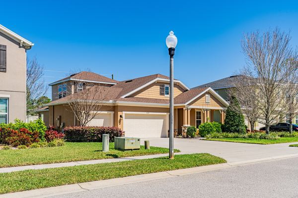 Angled front view of a tan stucco home with a shingle roof, a two-car garage and a covered entry, on a corner lot with a street lamp in the foreground.