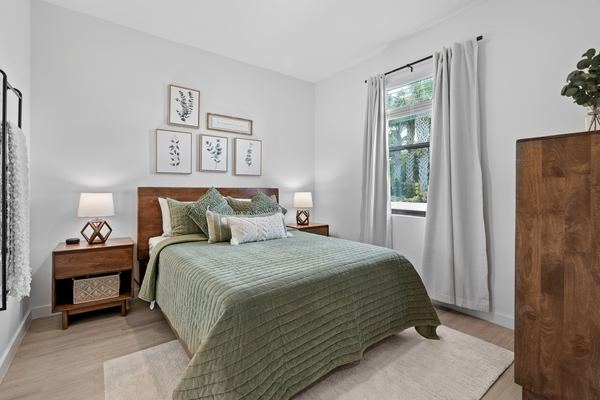 Bedroom with a wood headboard, a sage green quilt, matching bedside lamps, botanical prints above the bed and pale wood flooring.