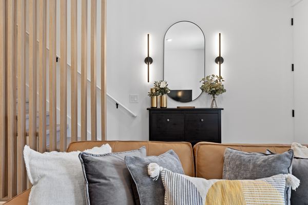 Arched mirror between two slim black sconces above a dark console, seen over a tan leather sofa, with a wood-slat stair screen at the left.